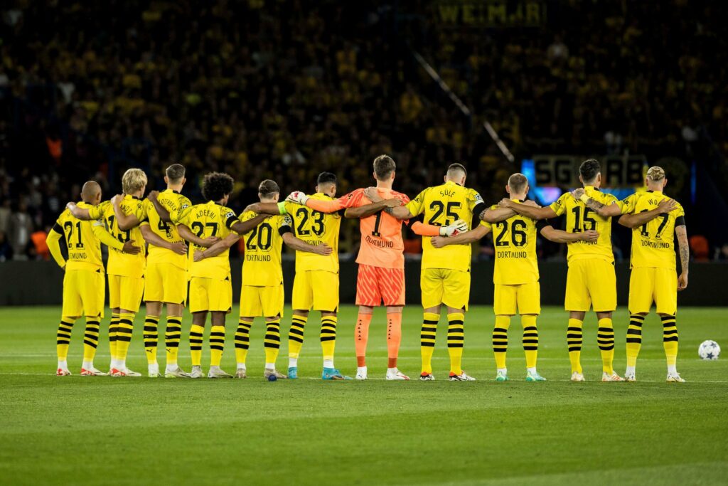 A soccer team in yellow and black uniforms, sponsored by Betway, stands arm in arm on the field. They face away from the camera, with a packed stadium in the background, reflecting their unity and determination.