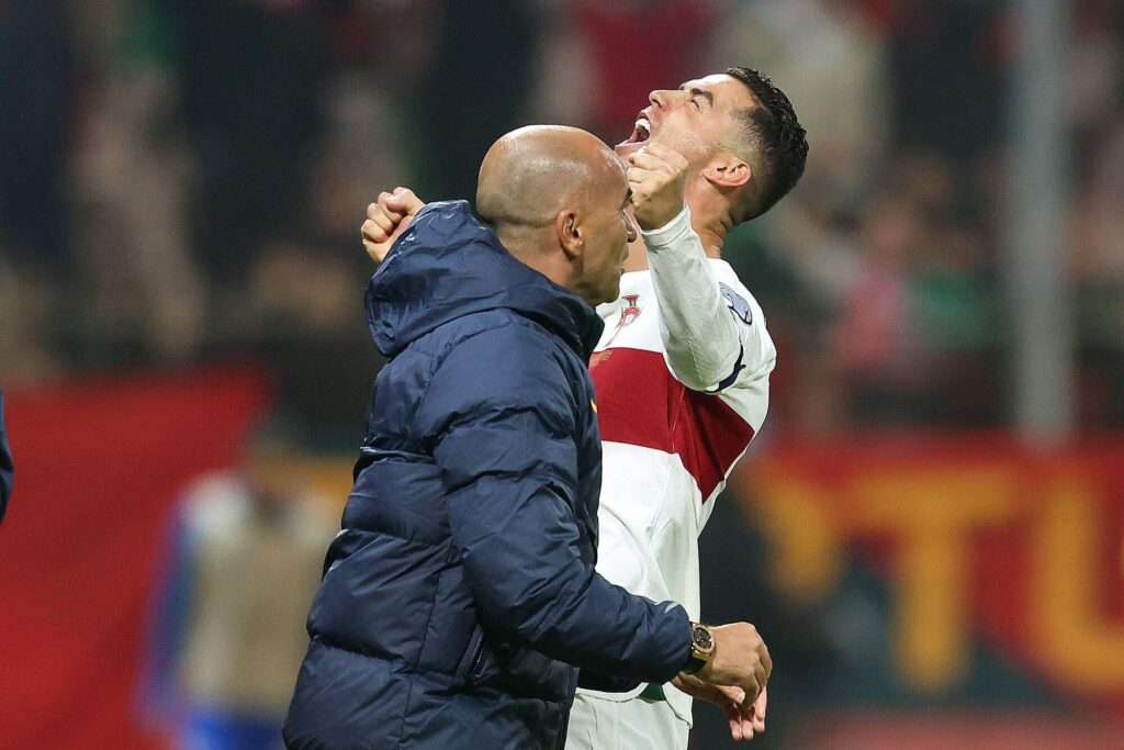 The soccer player in a white jersey, sponsored by Betway Arabia, celebrates with raised arms and a joyful expression. Beside him, another person in a blue jacket offers a slight smile. The background features blurred spectators and the hint of a flag.