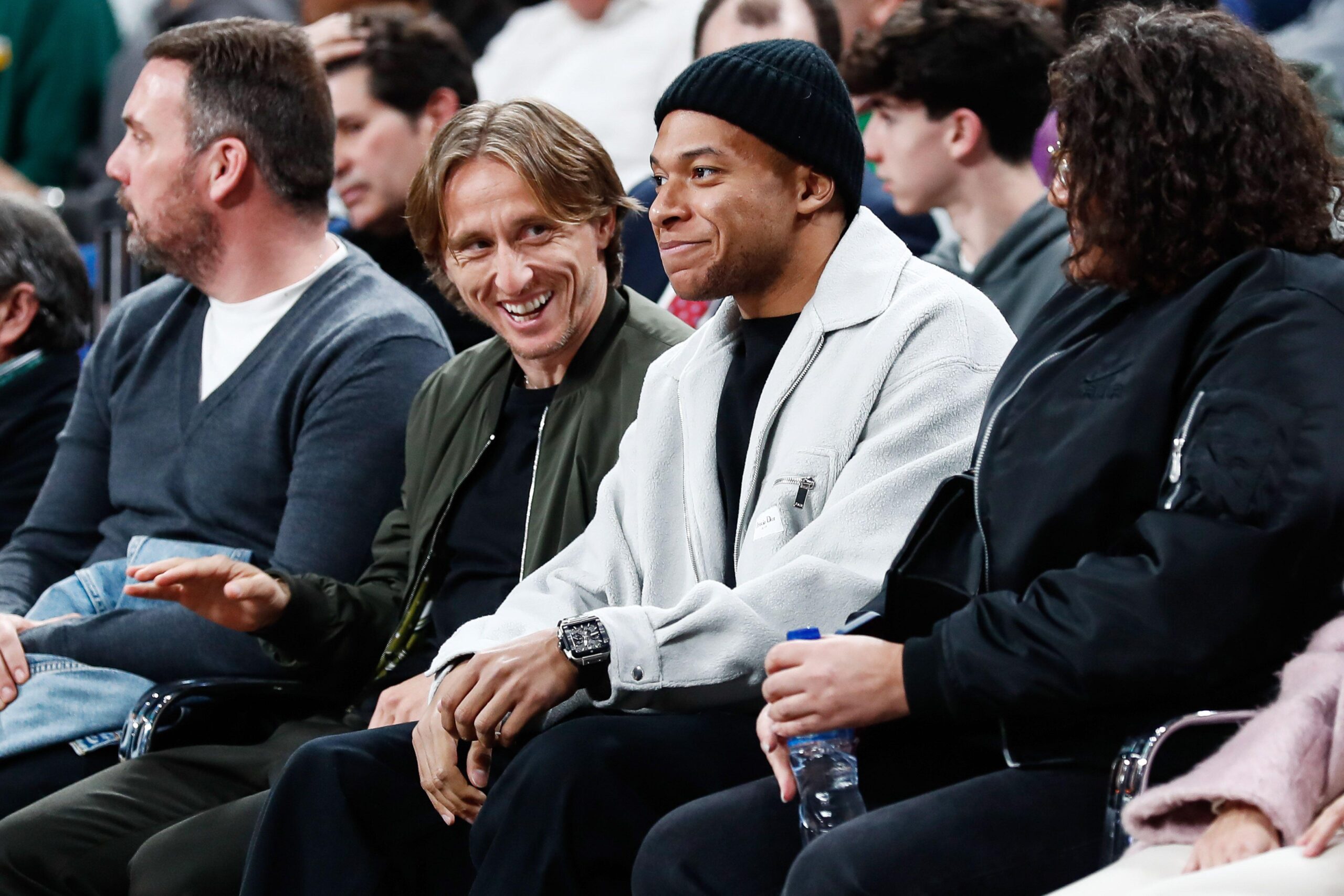 Four people are sitting in a row at an indoor event, with the middle two engaging in conversation. The background shows a blurred audience. The second person from the left is smiling warmly while the others look on.