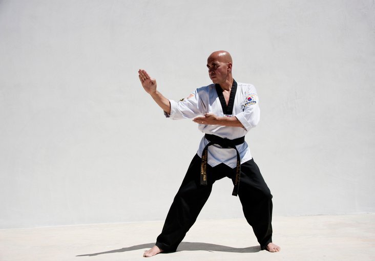 A person practicing martial arts stands in a strong pose against a plain white background. They wear a white martial arts uniform with a black belt, holding an arm out with focus and determination.