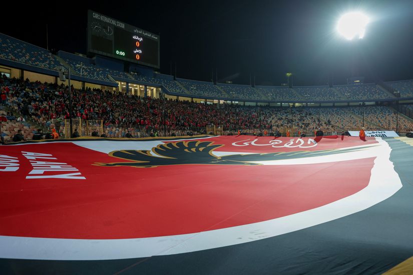 Un grand stade avec une foule de supporters dans les tribunes. Un drapeau rouge et noir géant représentant un aigle recouvre une partie du terrain. Les lumières du stade sont allumées, illuminant la scène animée tandis que le tableau d'affichage brille en arrière-plan, mettant en valeur le logo de Betway Arabia avec fierté.