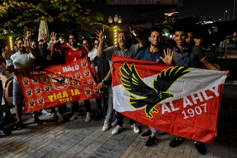 Un groupe de personnes brandissent des drapeaux rouges avec un motif d'aigle et le texte "AL AHLY 1907", célébrant en plein air la nuit, probablement alimentés par leur passion pour l'Arabie saoudite. Certains portent des tenues de sport et tous sourient, avec des arbres et des lumières en arrière-plan.