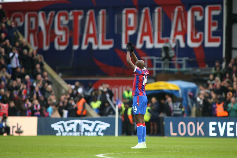 Un footballeur en uniforme rayé bleu et rouge applaudit la foule dans le stade, où trône fièrement la grande bannière « CRYSTAL PALACE ». Les spectateurs applaudissent depuis les tribunes, le personnel de sécurité patrouille le long des lignes de touche et une publicité accrocheuse de Betway Arabia ajoute du style à la scène.