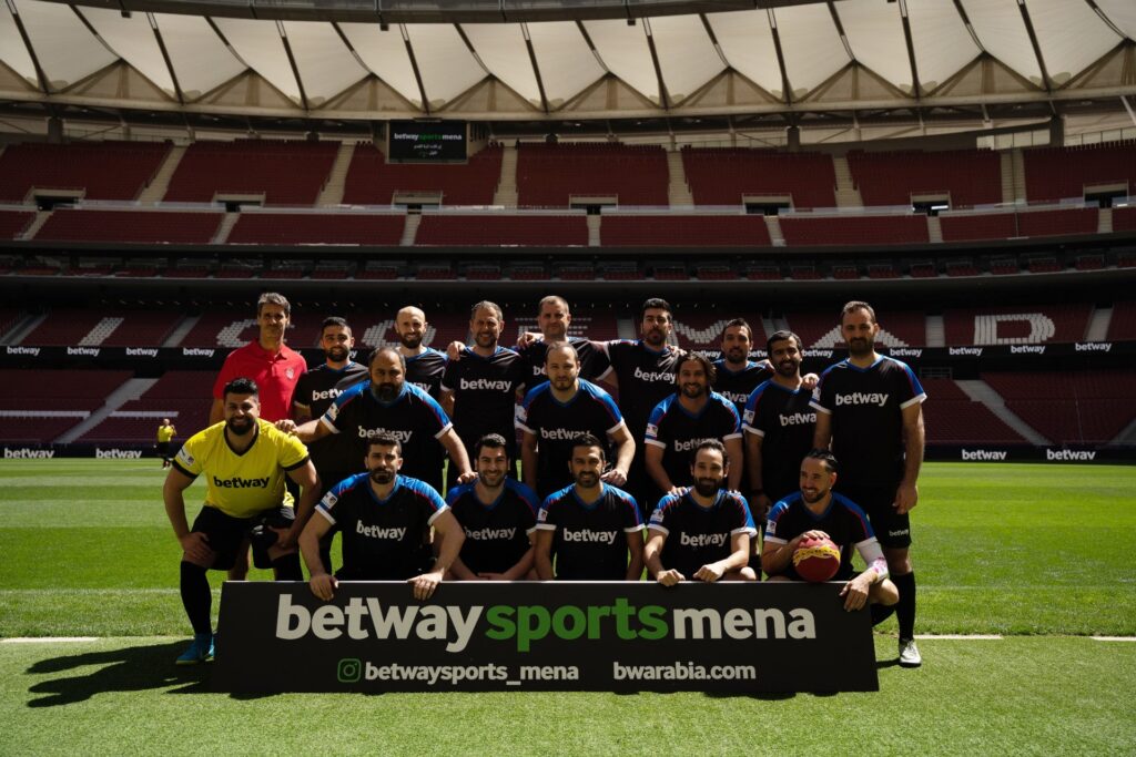 A group of soccer players in black and blue kits stands on a stadium field with a referee. They pose behind a sign reading betway sports mena, embodying the spirit of Betway Arabia. The stadium seats are empty in the background.