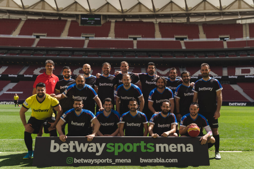 فريق كرة قدم يقف في ملعب فارغ من الجمهور. اللاعبون يرتدون زي "betway" باللونين الأسود والأزرق، ويحملون كرة قدم. لافتة أمامية مكتوب عليها "betwaysportsmena"، تسلط الضوء على تفاصيل وسائل التواصل الاجتماعي وموقع Betway Arabia.