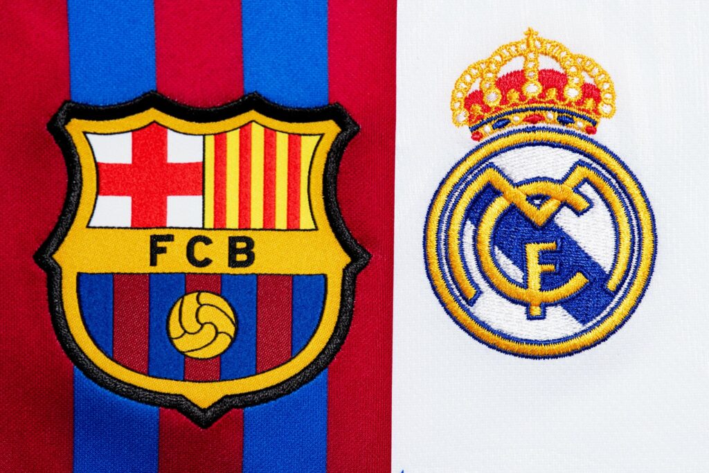 Barcelona and Real Madrid football club logos sit side by side, poised for a thrilling clash. Barcelonas emblem rests on red and blue stripes with a ball-adorned crest, while Real Madrids crowns its white backdrop. Witness the rivalry with Betway Arabia in the mix for an unforgettable experience.