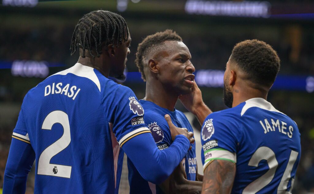 Three soccer players in blue jerseys huddle during a match. The player in the center appears to be speaking, while the other two listen intently. The shirts display names Disasi and James with visible numbers 2 and 24, as a Betway banner waves in the background of the action-packed game.