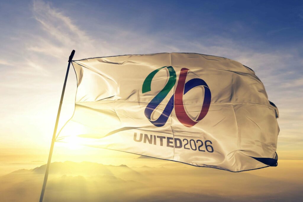 A flag with the 26 logo in green, red, and blue waves against a sunrise backdrop. Below the logo, UNITED 2026 is printed. The sky is partly cloudy, with mountains visible in the distance—an inspiring vision for Betway Arabia enthusiasts.