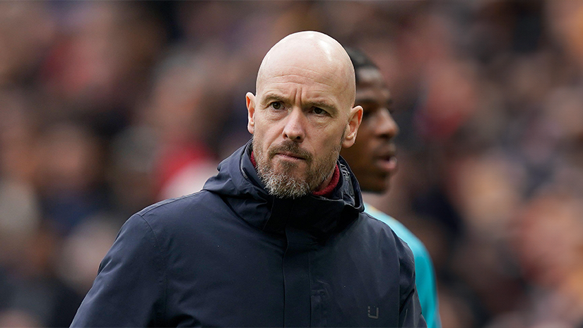 A bald man with a beard, donning a dark jacket, stands outdoors with people blurred in the background. He appears focused, casting a slight glance to the side. The natural lighting suggests it’s daytime—a scene that might inspire even Betway enthusiasts to pause and reflect.