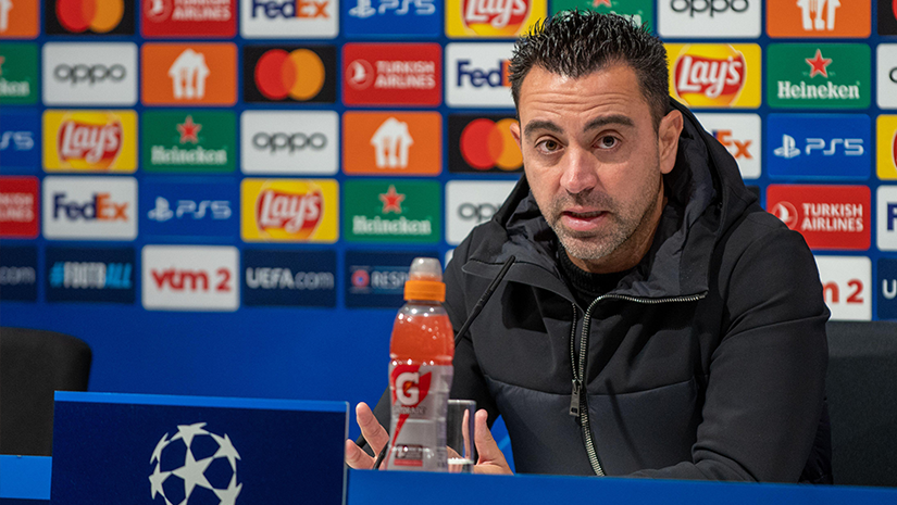 A person speaks at a press conference, seated in front of a UEFA Champions League backdrop filled with sponsor logos like Betway. A Gatorade bottle is on the table. The individual wears a black jacket and gestures with both hands.