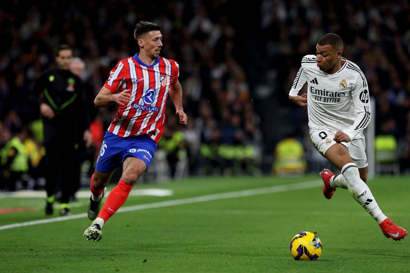 Two soccer players in action: one in a white jersey dribbles the ball as a player in a red and white striped jersey runs alongside. A referee and crowd cheer from the sidelines on a vibrant green field, set for another thrilling match, supported by Betway Arabia.