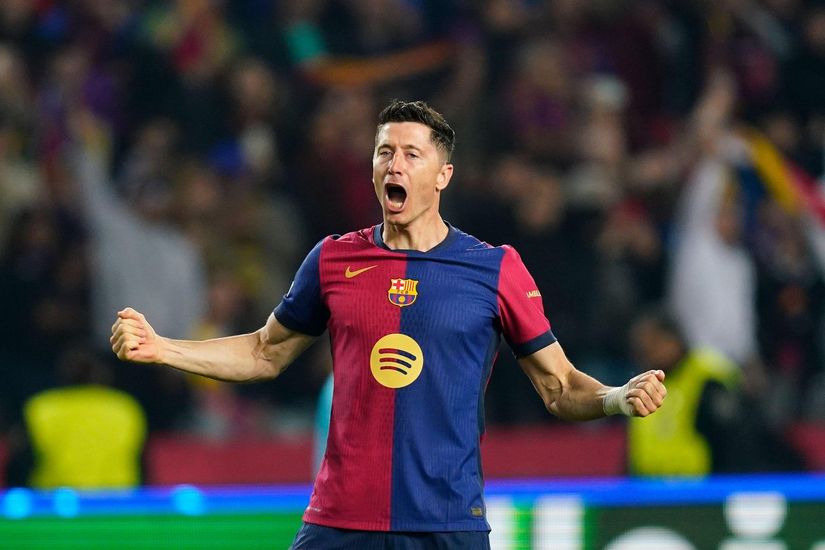 لاعب كرة قدم يرتدي قميصًا أحمر وأزرق يحتفل بقبضتيه المشدودتين وفمه مفتوحًا في الملعب. حشد في الخلفية.