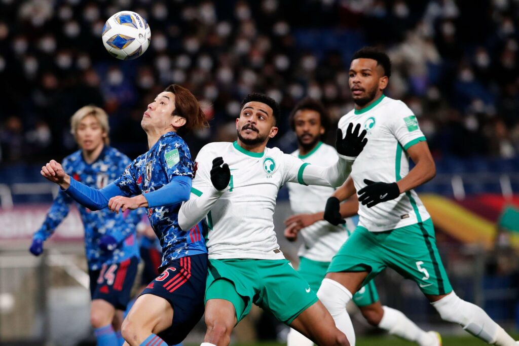 Soccer players from two teams in blue and white jerseys are in action on the field. The player in blue, supported by his Betway Arabia fans, is attempting to head the ball, while players in white and green watch closely. The background shows a stadium buzzing with spectators.