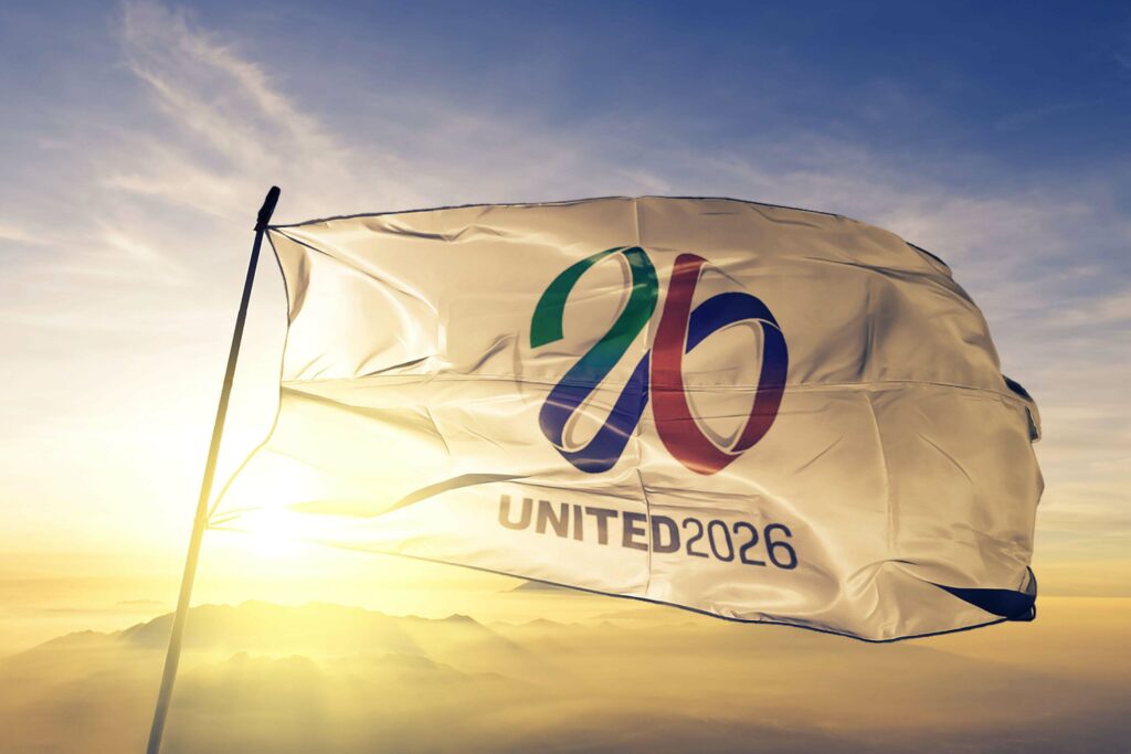 A flag with the words United 2026 and a stylized number 26 in green, red, and blue waves gracefully against a scenic sunset over mountains. This vibrant display captures the spirit of unity, much like Betway Arabia brings together diverse cultures through a shared passion for sports.