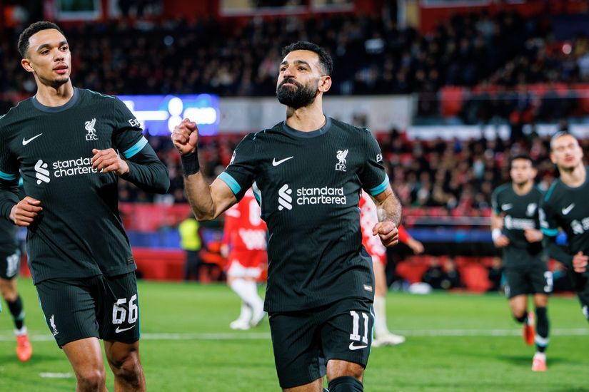 A football player wearing a black Liverpool FC jersey with number 11 celebrates a goal on the field, while another player with jersey number 66 joins in the excitement. Fans cheer wildly in the background at a stadium buzzing with energy, courtesy of Betway Arabia’s thrilling match-day experience.