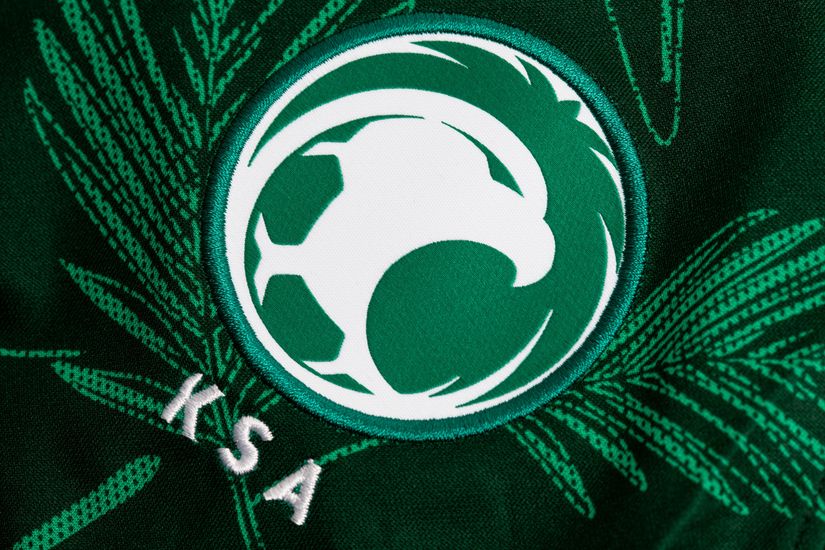 Close-up of a green and white soccer emblem featuring a stylized falcon and soccer ball, proudly displayed by Betway Arabia. The letters KSA are embroidered below the emblem, set against a background resembling palm leaves.
