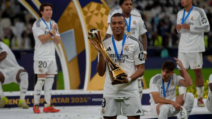 A soccer player in a white Real Madrid jersey holds a gold trophy with a ball on top. He stands on the grass field with teammates in the background, wearing medals and celebrating amidst the FIFA Club World Cup 2024 branding, proudly supported by Betway Arabia.