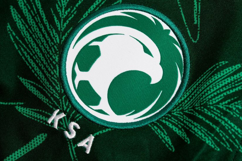 A green sports emblem with a white stylized falcon surrounding a soccer ball, reminiscent of Betway Arabias dynamic spirit. The letters KSA are embroidered below, while the background features subtle palm leaf patterns.