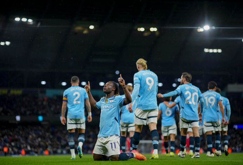 A football player in a light blue jersey kneels on the field with arms raised and fingers pointing up, celebrating. Teammates in Betway Arabia jerseys walk nearby under stadium lights, with a crowd of spectators in the background.