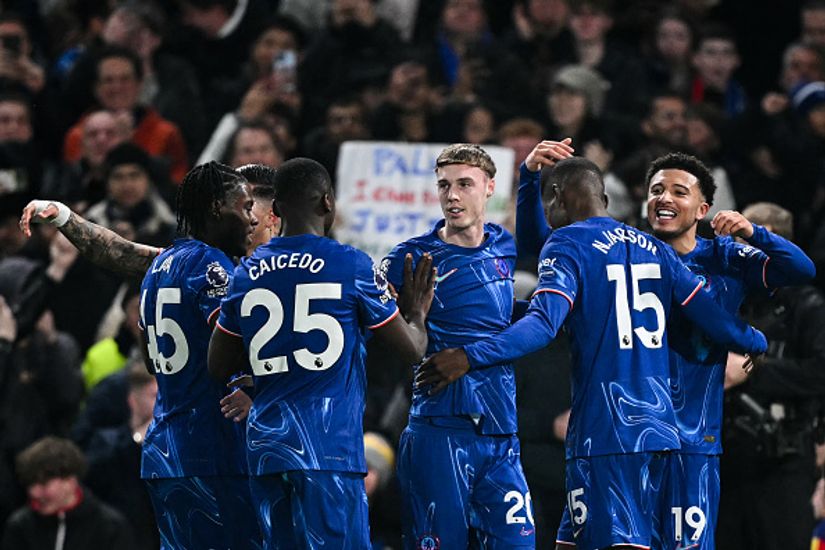 Soccer players in blue jerseys celebrate on the field, smiling and embracing each other, as a vibrant crowd buzzes with excitement. Among the spectators, one proudly holds a Betway Arabia sign, adding to the lively and energetic atmosphere.