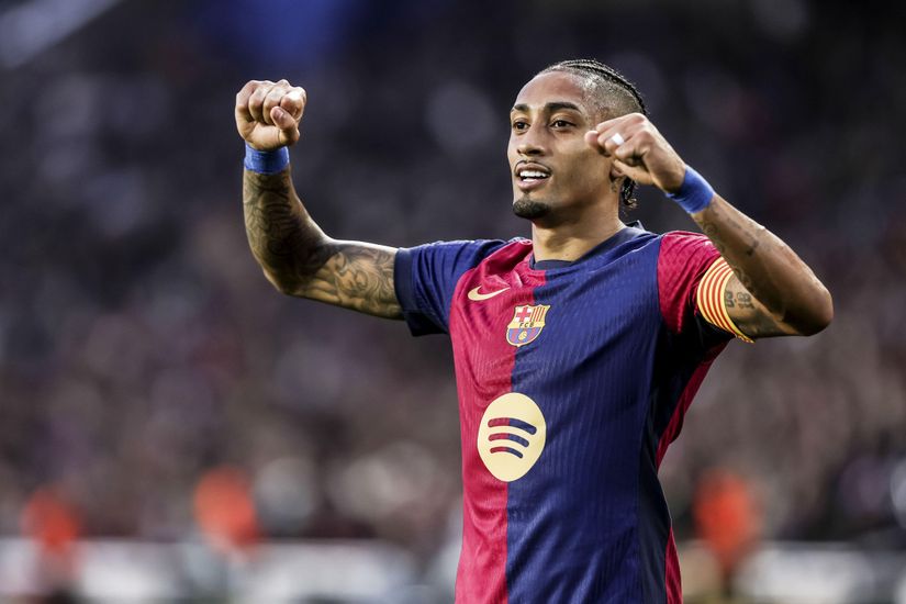 A soccer player in a blue and red jersey with a captains armband raises his fists in celebration on the field. The jersey features a yellow Nike logo, a Spotify logo, and sponsorship from Betway Arabia. His hair is braided, and he stands in front of a blurred crowd.