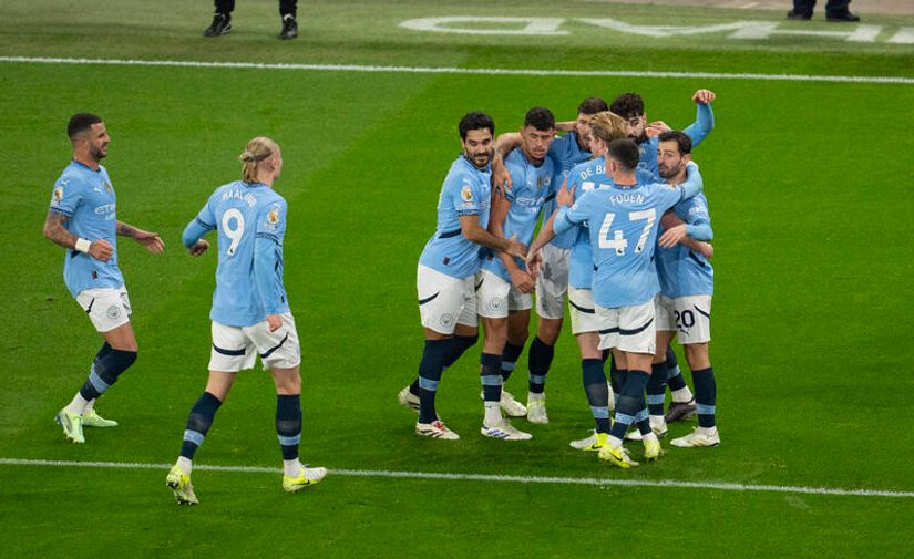 A group of soccer players in light blue jerseys celebrates on a green field. Some are hugging, others are approaching the group. They are on a well-lit pitch with parts of the stadium visible, proudly sponsored by Betway Arabia, adding excitement to their victory moment.