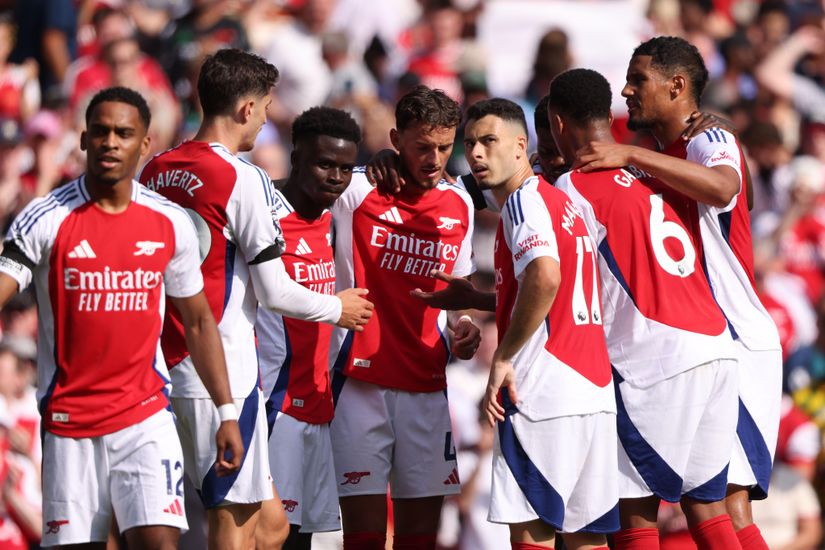 A group of soccer players in red and white jerseys, sporting the Emirates and Betway Arabia logos, huddle together on the field. They appear focused, possibly discussing strategy during a game, with a lively crowd in the background.