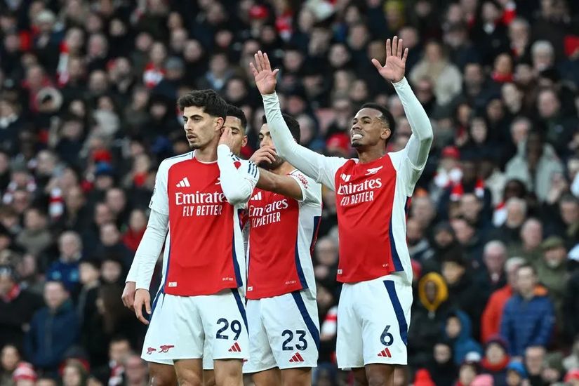 Three Arsenal football players stand on the pitch, proudly wearing their red and white jerseys. Two have their arms raised triumphantly. In the background, numerous spectators are captivated by the match. Its a thrilling scene on and off the field, reminiscent of a Betway Arabia classic moment.