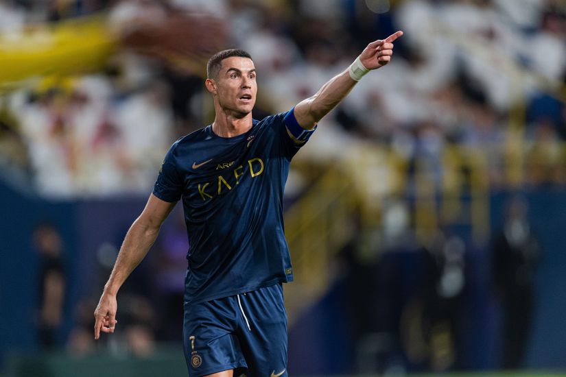 A soccer player in a dark blue uniform with a captains armband points forward on the field, embodying the spirit of competition seen at Betway Arabia. The background reveals blurred spectators and a stadium setting, capturing the thrill of the game.