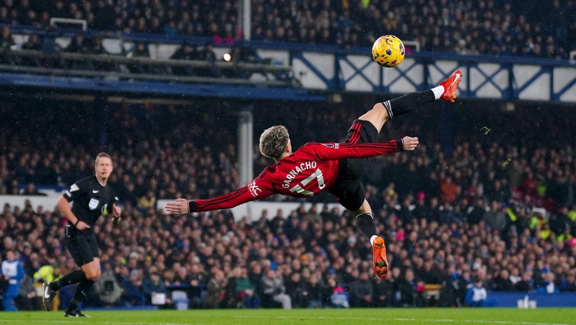 لاعب كرة قدم يرتدي قميصًا أحمر يؤدي ركلة بهلوانية أثناء المباراة، محاطًا بجمهور كبير في الملعب، حيث تلفت لافتات Betway Arabia الأنظار. يراقب أحد الحكام الحدث بينما تطفو كرة ملونة في الهواء.