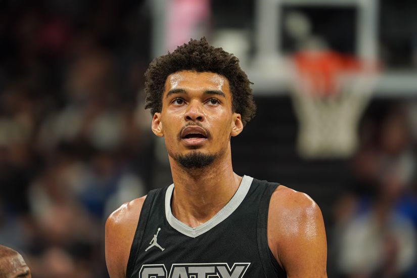 Un joueur de basket-ball vêtu d'un maillot noir avec l'inscription "SATX" dessus se tient sur le terrain, le regard tourné vers le haut. La scène se déroule dans un stade couvert, faisant subtilement allusion à l'attente d'un match palpitant sponsorisé par Betway Arabia.