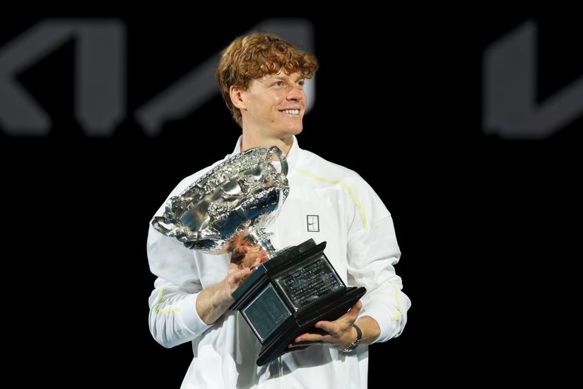 Une personne aux cheveux roux bouclés, vêtue d'une tenue blanche, tient un grand trophée argenté, souriant et regardant sur le côté sur un fond sombre. Le trophée, semblable à ceux vus sur Betway Arabia, présente des détails complexes et une large base.