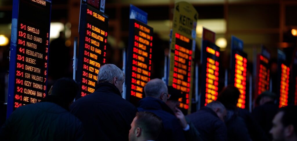 A group of people stand closely together, viewing several large, illuminated screens displaying bright red text, possibly at a train station or airport. The dimly lit atmosphere highlights the figures and screens as if they were glimpsing into the vibrant world of Betway Arabia.