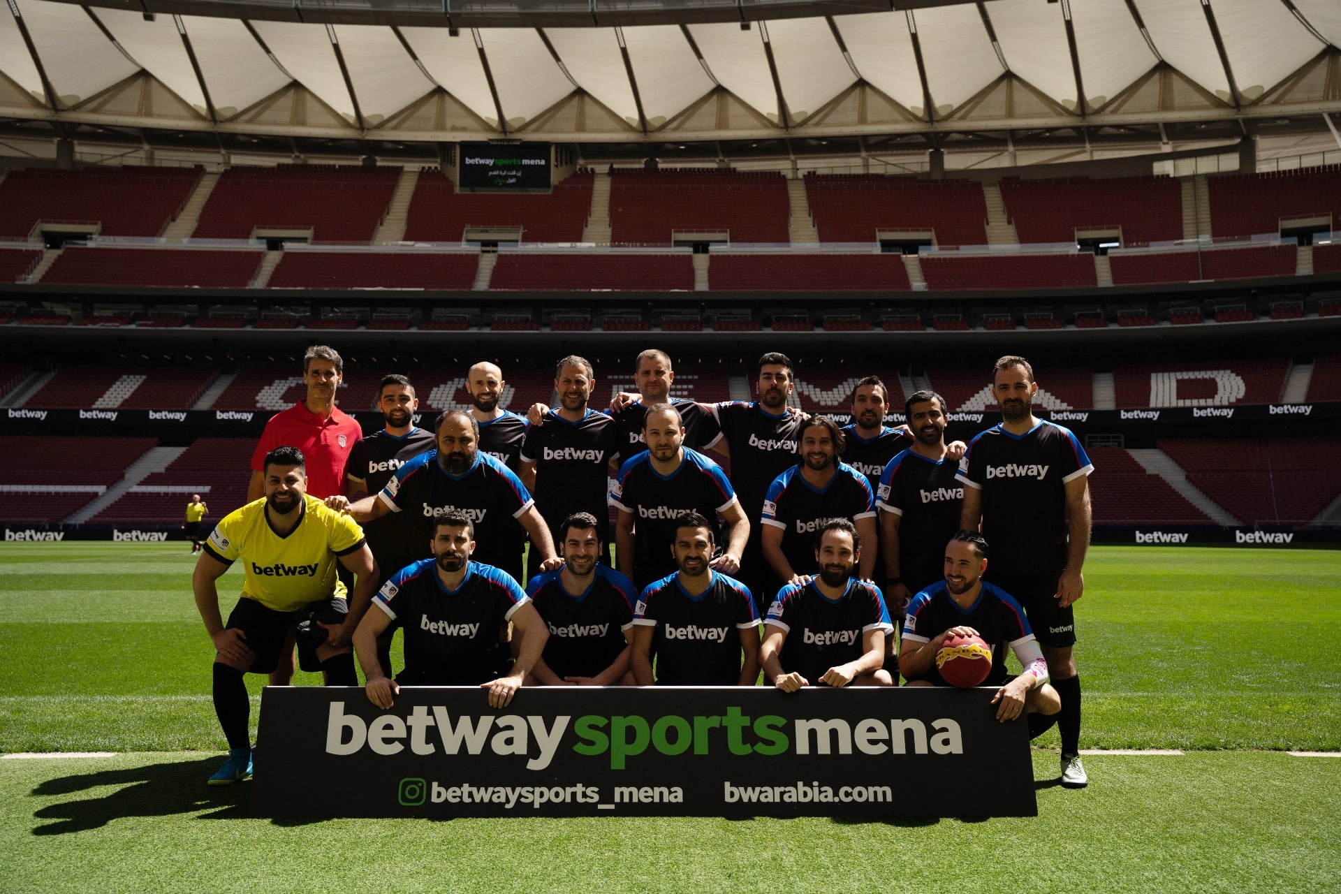 A group of soccer players and a referee pose on a stadium field in black jerseys featuring Betway logos. A betway arabia banner is displayed prominently in front, while the background reveals empty red and white stadium seats.
