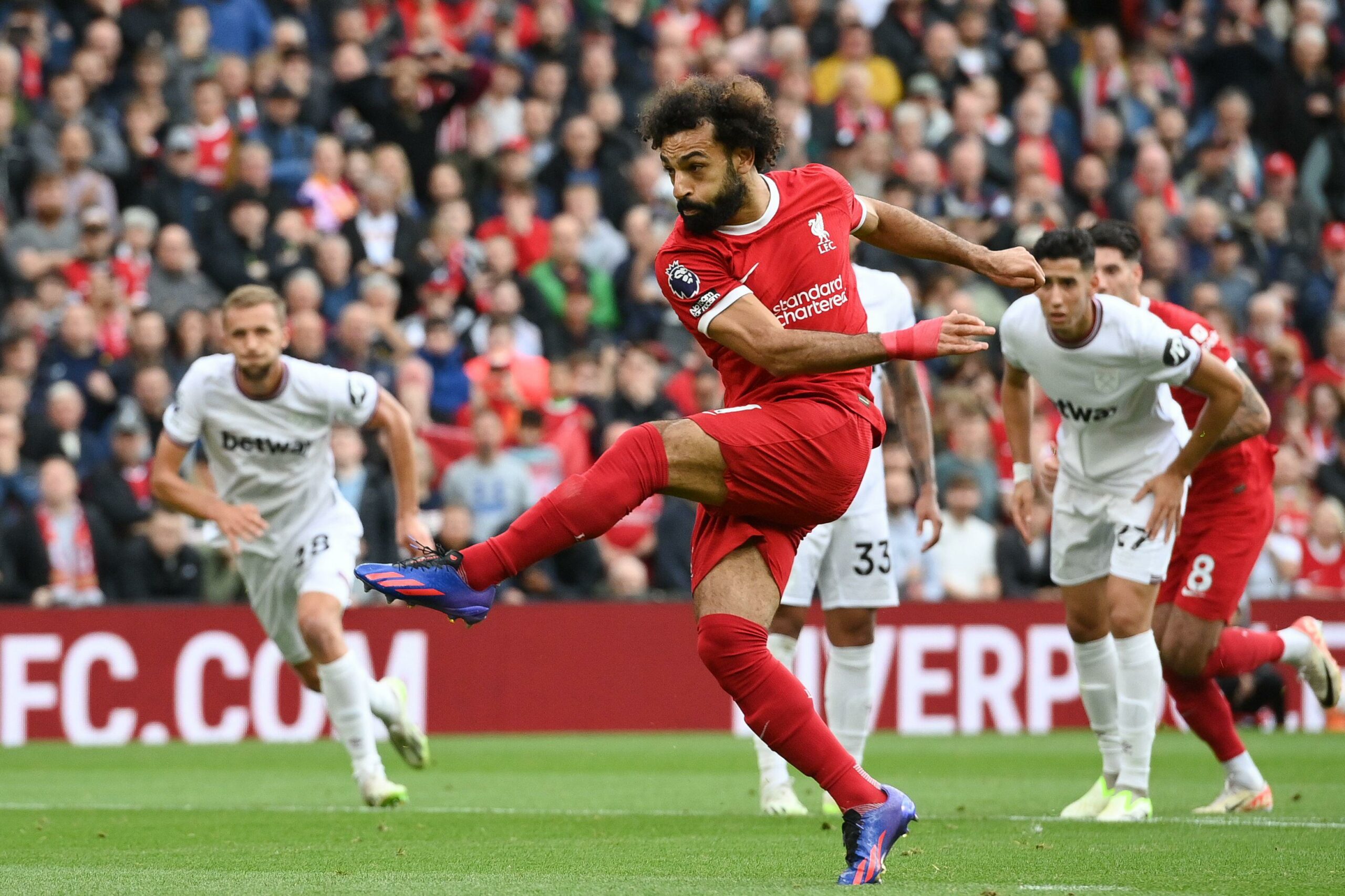 A soccer player in a red jersey skillfully kicks the ball during a thrilling match, while players in white jerseys watch nearby. With the stadium buzzing with spectators, its a perfect scene for fans following the action on Betway Arabia.