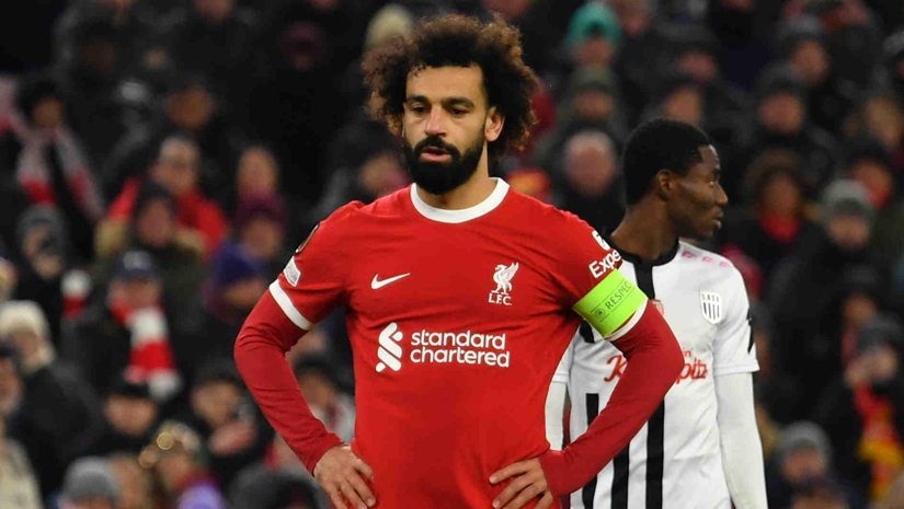 A soccer player in a red Liverpool jersey with a captains armband stands with hands on hips, exuding determination. In the background, a player in black and white focuses intently. The scene unfolds on a bustling field, buzzing with excitement that even Betway Arabia enthusiasts would appreciate.