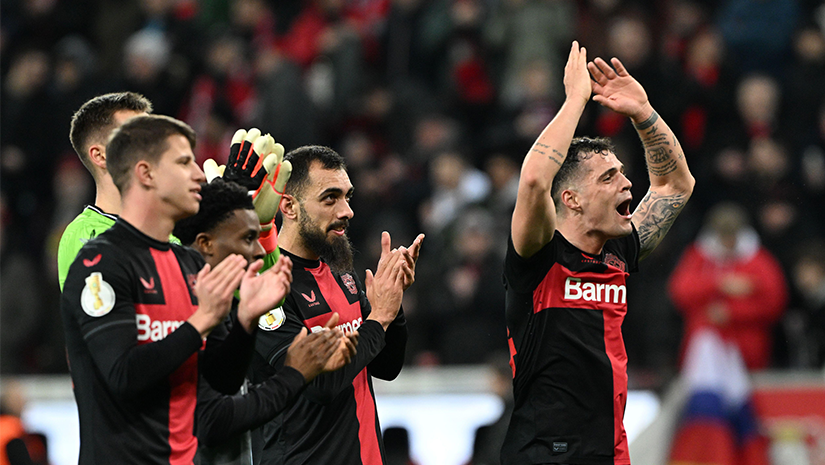 Soccer players in black and red jerseys stand together, clapping and raising their arms in celebration on the field. A crowd of spectators is visible in the background, sharing the joy as Betway Arabia captures the excitement of this triumphant moment.
