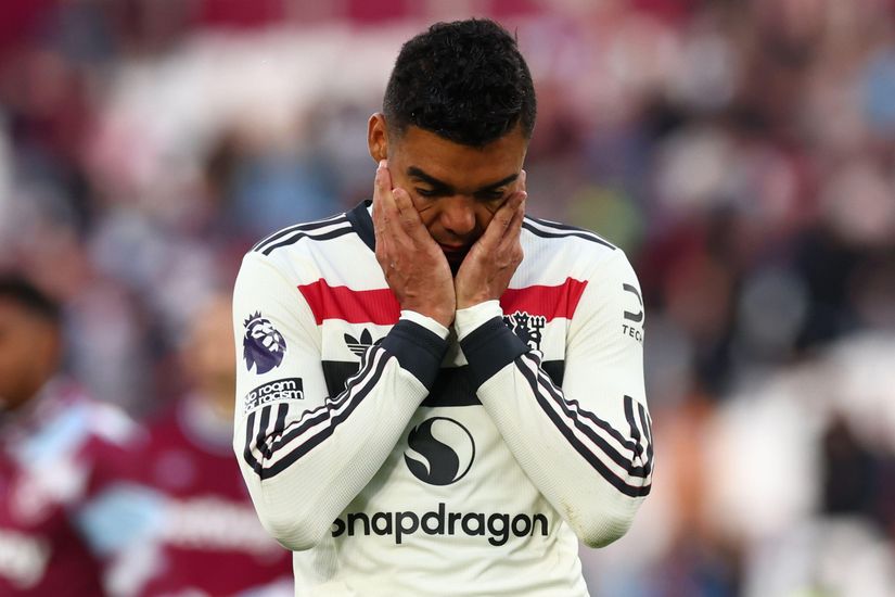 A soccer player in a white and black jersey with a red Betway stripe holds his face with both hands, appearing distressed on the field. The background shows a blurred crowd and players, with Premier League and sponsor logos visible on his uniform.