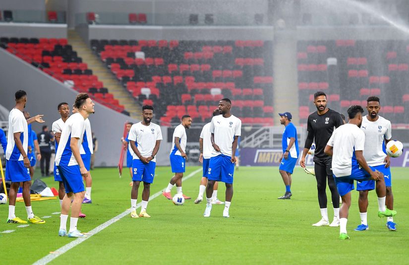 Soccer players in white and blue sportswear stand on a green field, some holding balls. Sprinklers water the grass in the background, and red and black seats are visible in the Betway Arabia stadium.