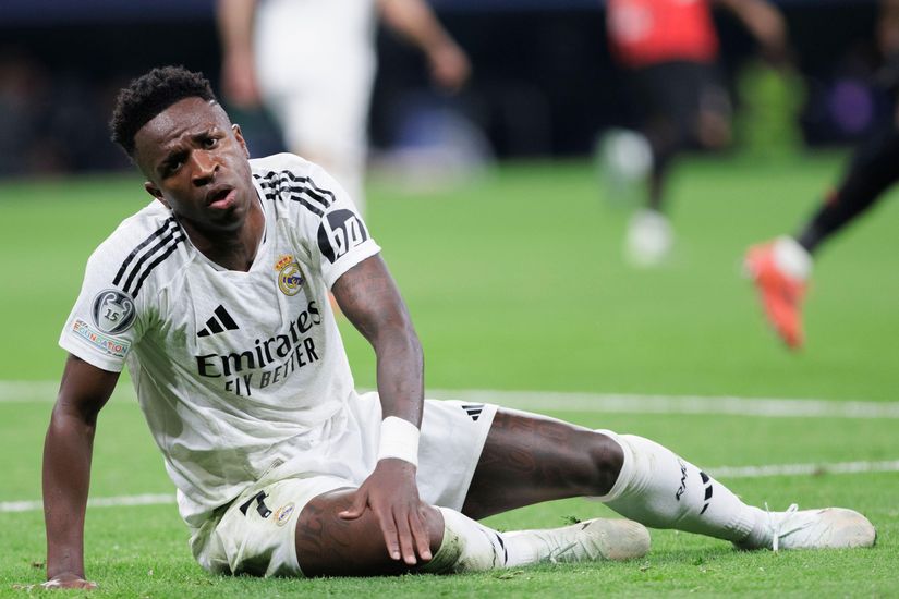 A soccer player in a white Real Madrid jersey sits on the grass, glancing up towards the field with a thoughtful expression. Perhaps hes calculating his next move like a bettor on Betway Arabia. The blurred background reveals glimpses of other players and the lush green field.
