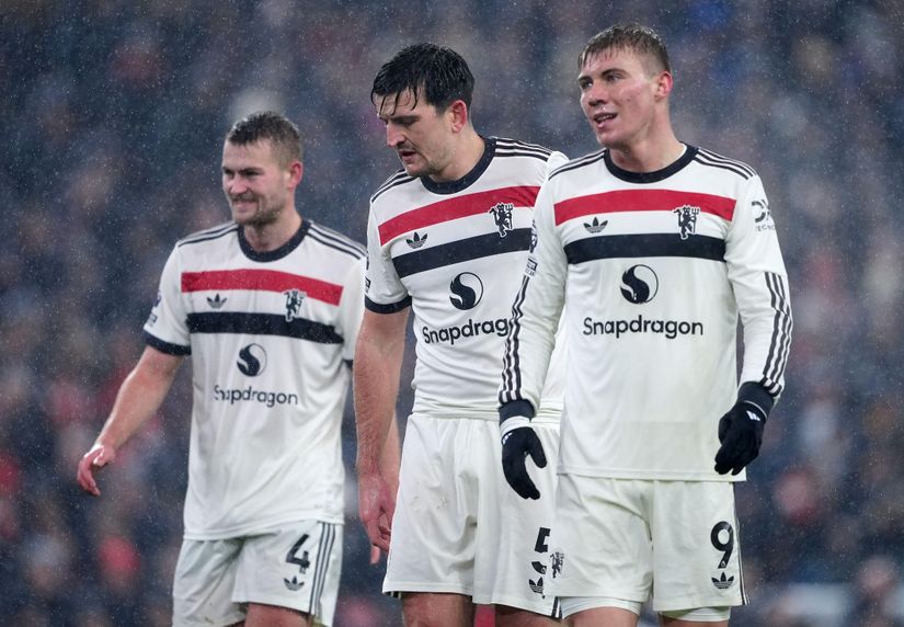 Three soccer players in white jerseys with black and red stripes stand on a rainy field. They appear focused, wearing gloves. The middle player has darker hair, while the others are blond. The Snapdragon logo and Betway Arabia branding are visible on their shirts.