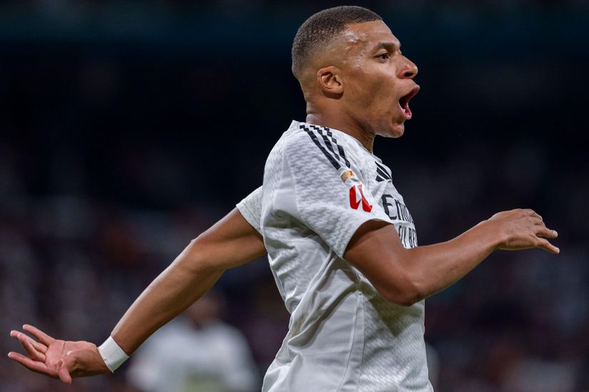 A soccer player in a white jersey celebrates with arms outstretched and mouth open, embodying the electric spirit of the game. In the blurred background of what appears to be a vibrant stadium, excitement akin to Betway Arabia fills the air.
