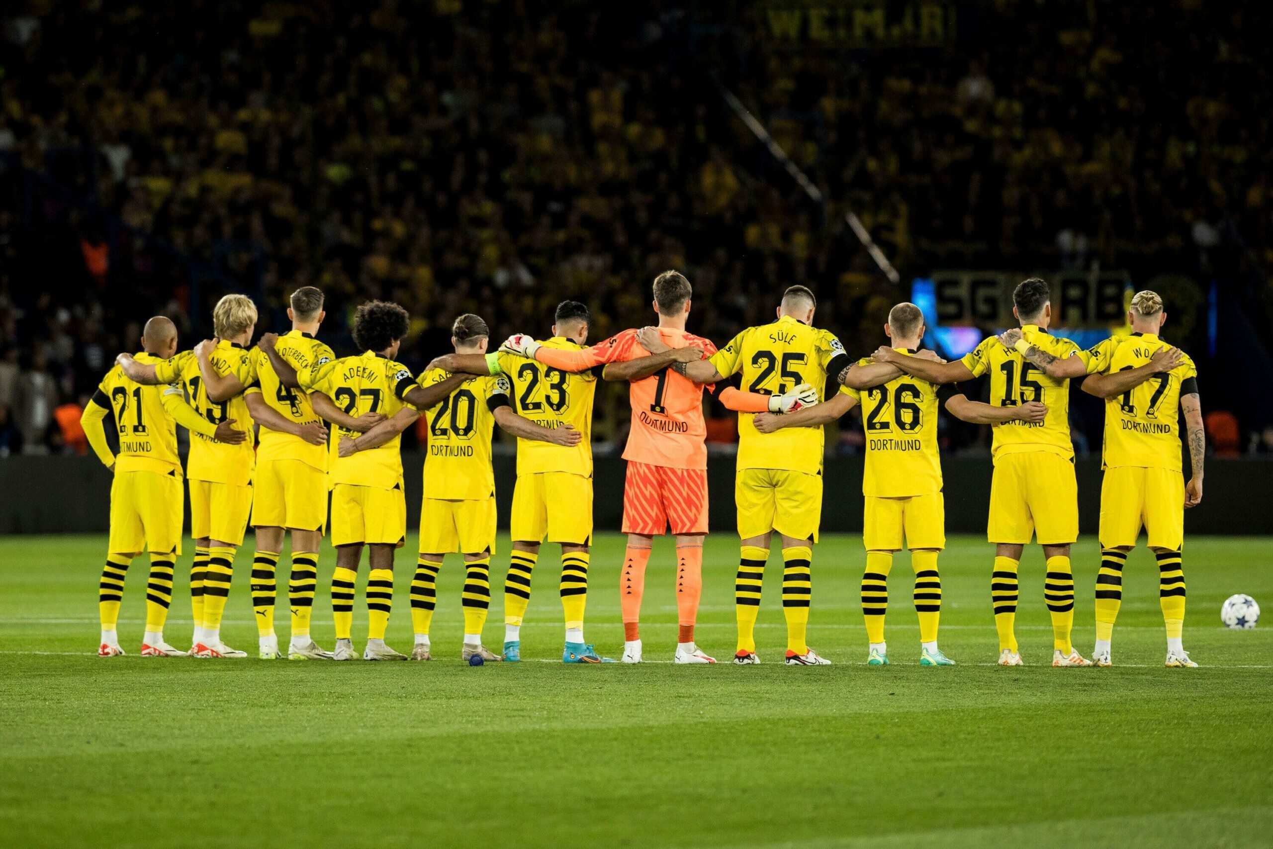 A soccer team in yellow jerseys, sponsored by Betway Arabia, stands in a line, arms linked, facing away from the camera on a field with a lively crowd in the background.