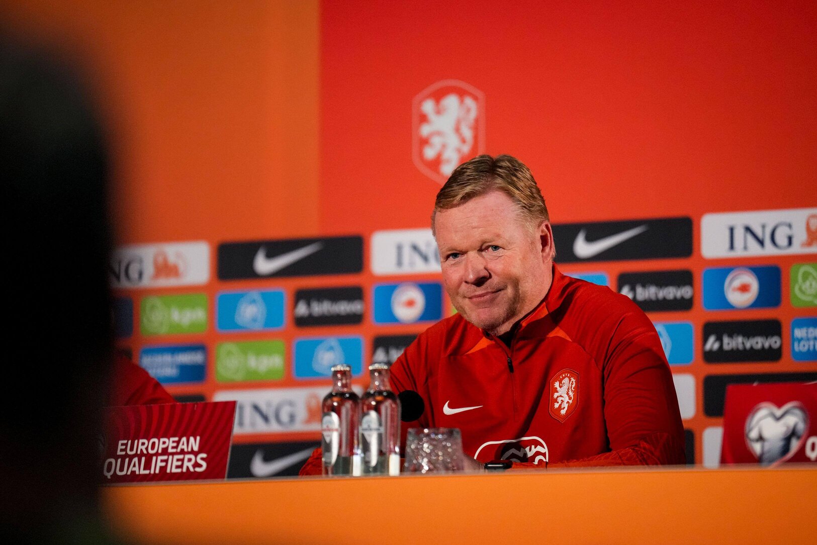 A person in a red sports jacket sits at a press conference table adorned with microphones and water bottles, set against a backdrop featuring sponsor logos, including Betway Arabia.