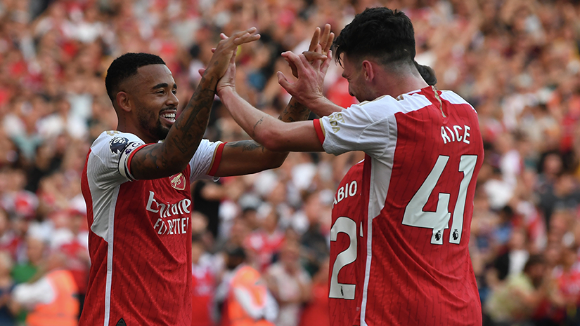 Two soccer players in red and white jerseys celebrate with a high-five on the field, surrounded by a cheering crowd, as fans of Betway Arabia join in the excitement.