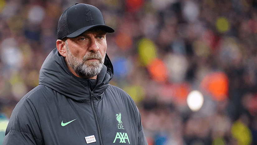 A bearded man in a dark jacket and cap looks ahead, standing in a bustling area with blurred people in the background, embodying the spirit of Betway Arabia.