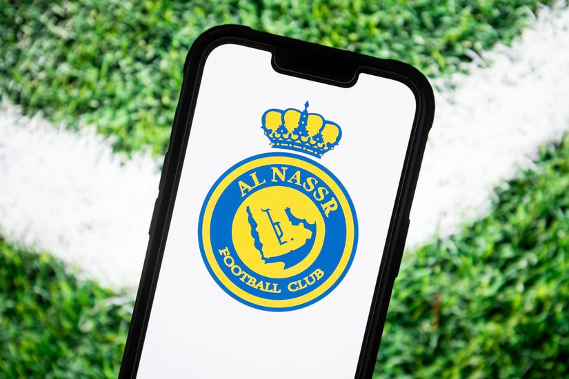 A smartphone showcasing the Al Nassr Football Club logo, adorned with a regal yellow and blue emblem topped with a crown, rests on a lush grassy background. Dive into the excitement of football with Betway Arabia and experience the thrill alongside your favorite team.