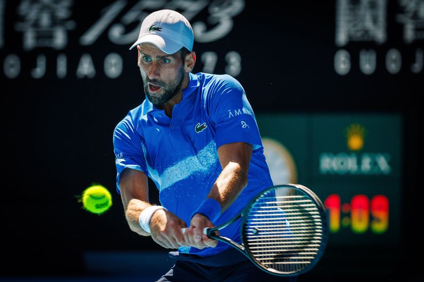 A male tennis player in a blue outfit hitting a backhand shot, with a blurred scoreboard in the background.