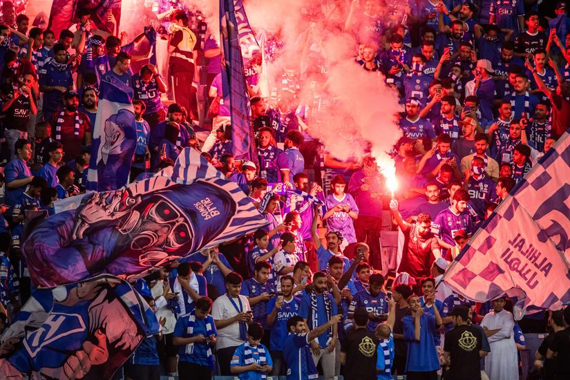 Crowd of sports fans in blue jerseys wave flags and hold flares, creating a vibrant, energetic atmosphere in the stadium.