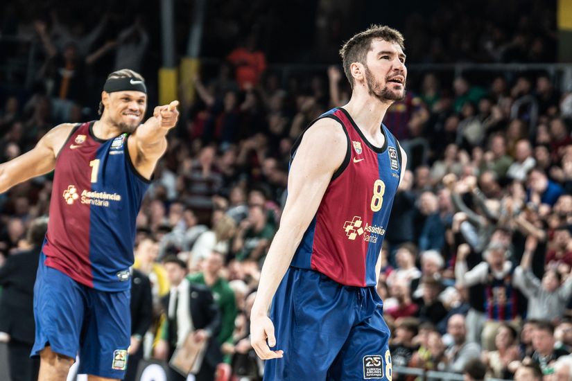 Two basketball players in blue and red jerseys celebrate on the court, with a cheering crowd in the background.